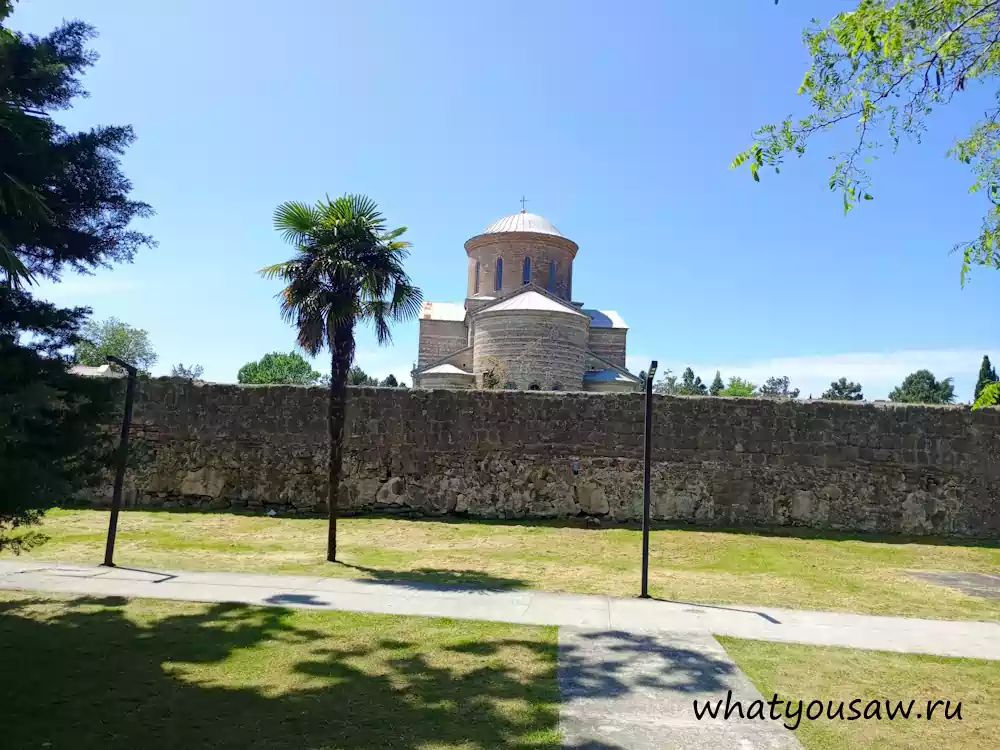 Вид с тропинки парка на храм, величественный каменный забор храма, ростущие в парке пальмы и сосны